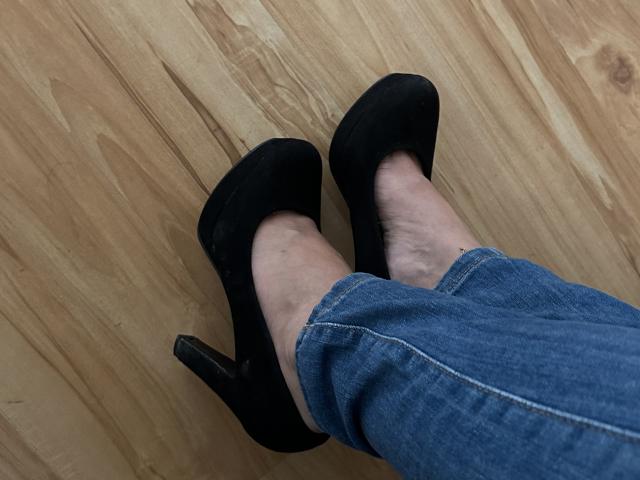 Stefanni's legs in blue jeans, wearing black high-heeled shoes, standing on a wooden floor. Her feet are slightly apart, showing the heels' pointed toes.