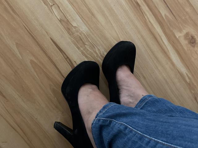 Stefanni's legs in blue denim, wearing black high-heeled shoes, standing on wooden floor. Her ankles and shoes are visible, with her legs slightly crossed.