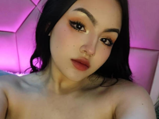 HaleiBeckham com cabelo escuro, delineador marcante, sombra laranja, lábios vermelhos cheios, pele clara, nariz pequeno, sem blusa, fundo rosa-roxo, olhando para a câmera, iluminação suave..