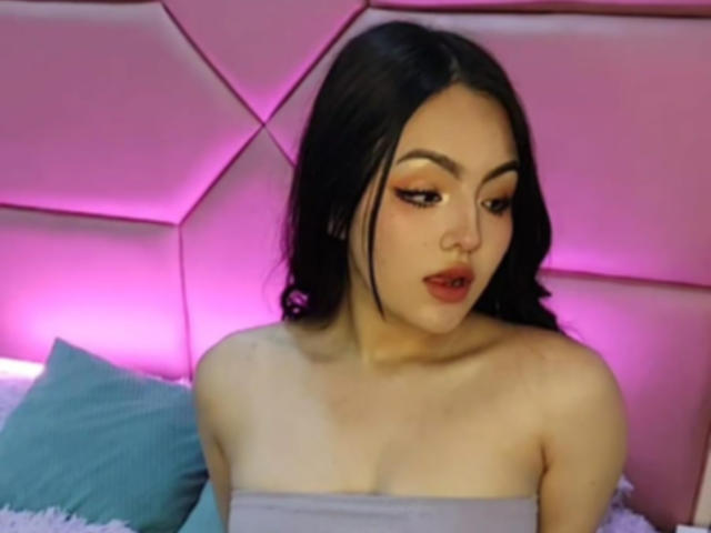 Halei Beckham mit dunklem Haar, Make-up und rotem Lippenstift, die leicht schmollend nach unten schaut, trägt ein graues Top ohne Träger, das ihre Brüste zeigt, vor einer rosa, gepolsterten Wand..