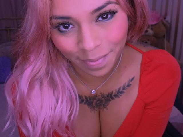 Chili, with pink hair and red top, smiling while leaning forward, showing her large cleavage and black floral chest tattoo.