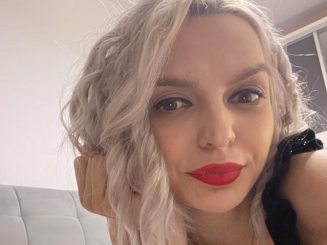 DouceAmelia with wavy silver hair, red lipstick, and black lace top, looking at the camera with a slight smile, her nose ring visible.
