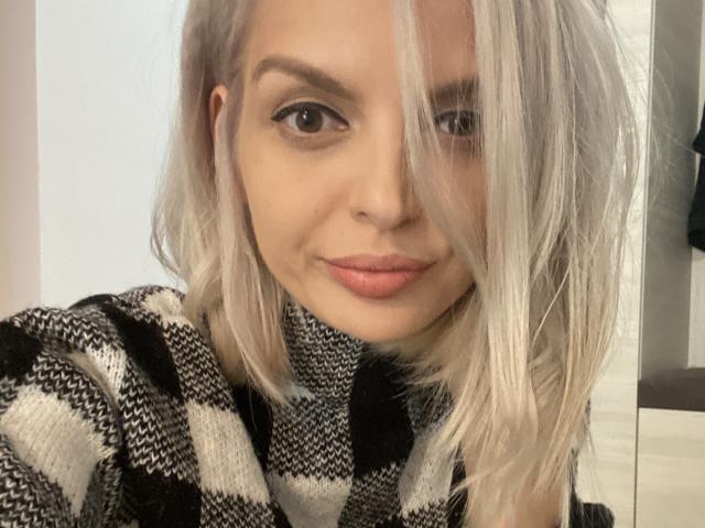 DouceAmelia with platinum blonde hair, black and white checkered sweater, full lips, and subtle makeup, looking directly at the camera with a slight smile.