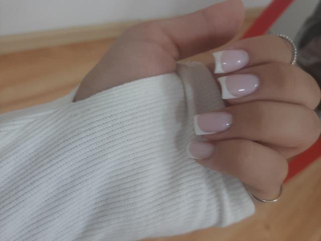 Destinis' hand with white ribbed sweater, pale pink French-manicured nails, silver ring, and beige hair tie, on a wooden floor.