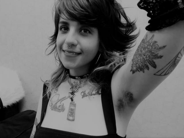 AmySpider with medium tits, tattoos, and arm hair, smiling, raising her right arm, wearing a choker and lace glove, in a black-and-white selfie.