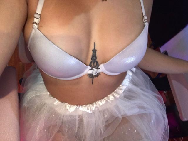 CamilaColeman's tits in a white bra, showing a black tattoo between her boobs, wearing a white tulle skirt. Her tan skin is visible, with her arms resting.