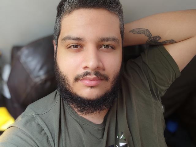 JeremmyJizz, un uomo corpulento con una barba e un baffo grigi, indossa una camicia verde, alza un braccio per mostrare un tatuaggio sul bicipite, e guarda la telecamera..