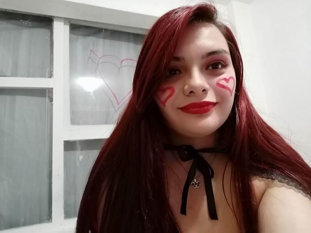 KatherinLatin with red lipstick, red face paint, and red highlights in her long hair, wearing a black choker, smiling at the camera. Her tits are partially visible.
