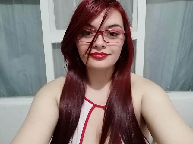 KatherinLatin with red hair, glasses, red lipstick, and white halter top with red straps, showing her boobs, sitting in front of a window.