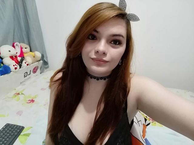 KatherinLatin with long brown hair, black choker, and black bow, wearing a low-cut black top, showing her small tits, smiling, in a bedroom with stuffed animals.