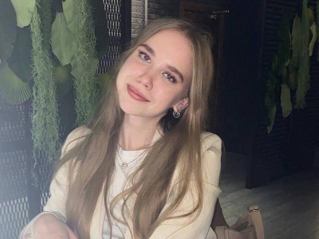 BunnyNiki, com longos cabelos loiros, sorrindo suavemente, olhos azuis, vestindo uma blusa branca, seios pequenos, maquiagem discreta e um colar delicado, sentada em um quarto com pouca luz..