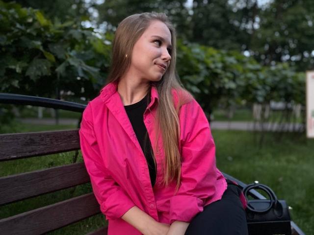 BunnyNiki, com cabelos castanhos longos, uma camisa rosa e calças pretas, está sentada em um banco de madeira, sorrindo de lado, com as mãos no colo, e uma bolsa preta à direita..