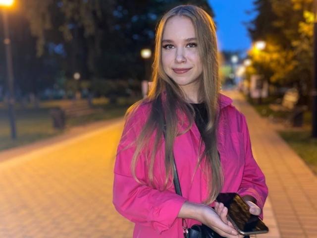 BunnyNiki mit langen blonden Haaren, einer pinken Jacke, einem schwarzen T-Shirt, die ein schwarzes Telefon hält, steht auf einem mit Ziegeln gepflasterten Weg in der Dämmerung, lächelt sanft, und im Hintergrund ist ein blauer Himmel zu sehen..