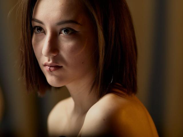 AlexaJang heeft kort bruin haar, subtiele make-up, en is topless waarbij haar borsten licht zichtbaar zijn. Ze ziet er serieus uit, met een zacht, natuurlijk licht op haar gezicht..