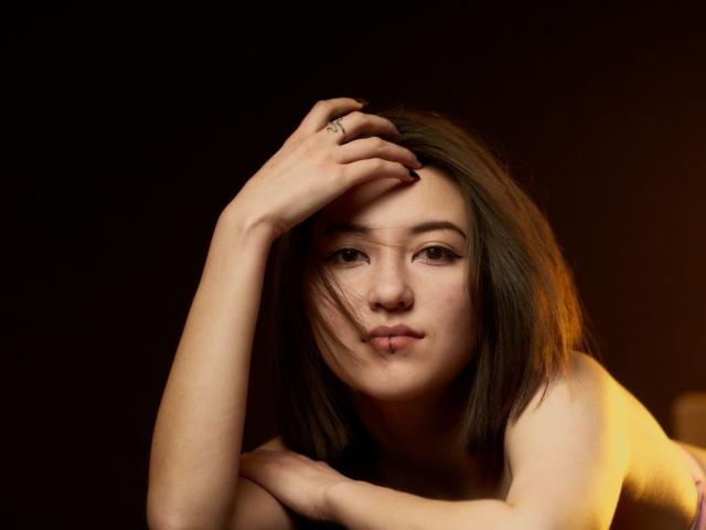 AlexaJang, con cabello castaño, labios carnosos y piel clara, lleva un anillo en su mano derecha, con la cabeza apoyada en su brazo izquierdo, sobre un fondo oscuro..