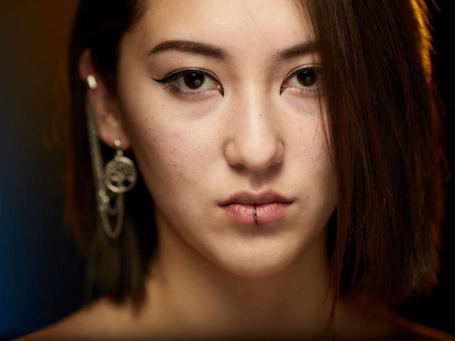 AlexaJang's face with dark brown eyes, winged eyeliner, short brown hair, nose piercing, skull earrings, and a small lip piercing. Her skin is light with a few blemishes.