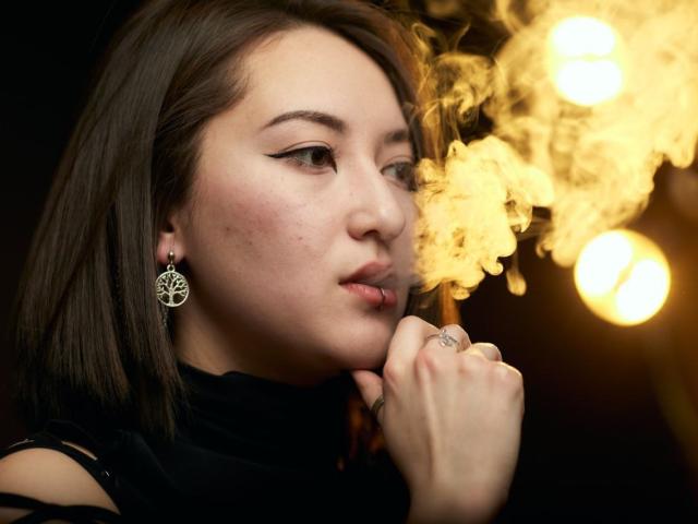 AlexaJang with smoky breath, brown hair, black turtleneck, winged eyeliner, pentagram earrings, ringed finger, and golden light, looking off to the side.