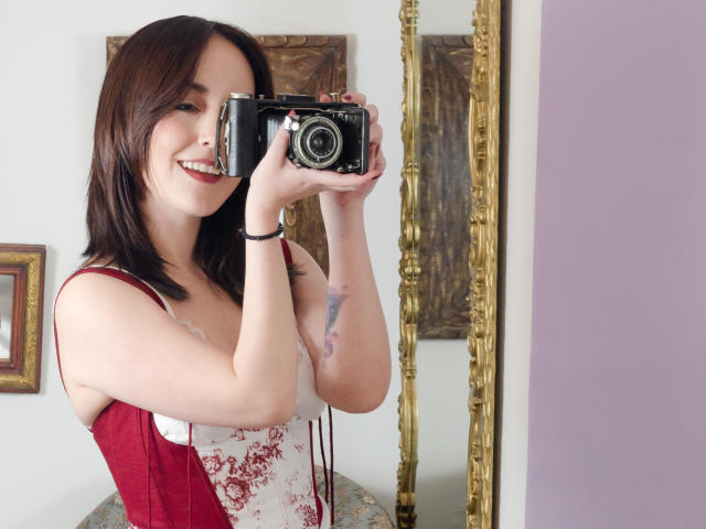 MollyMarmalade with shoulder-length brown hair, smiling, holding a vintage camera, wearing a red and white floral dress, visible tattoo on her right arm, standing in front of a mirror.