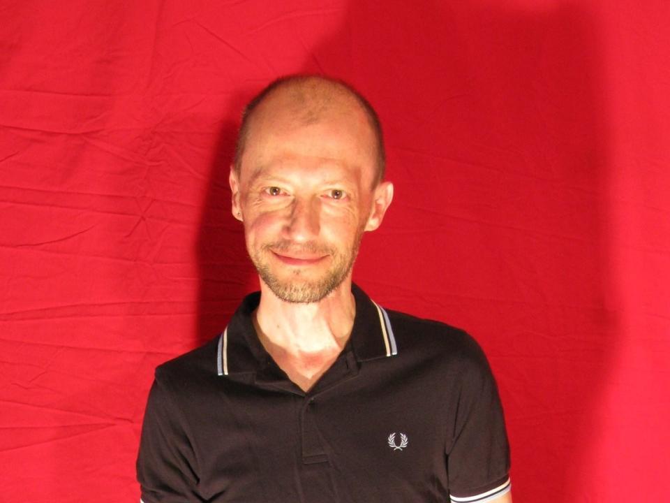 SilvainBoby69, calvo, sorridente, indossa una polo nera con bordi bianchi, con una leggera barba, sfondo rosso, corporatura media, aspetto casual, sicuro di sé, pelle chiara, piccolo logo sulla maglietta..