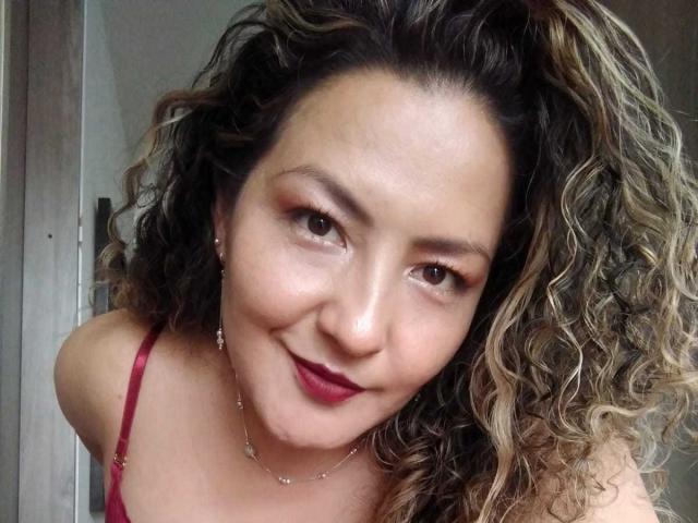 Amandabella69 ha i capelli castano ricci, rossetto rosso e un sorriso carino, indossa un reggiseno rosso, mostrando il seno, e guarda la fotocamera con gli occhi castani..