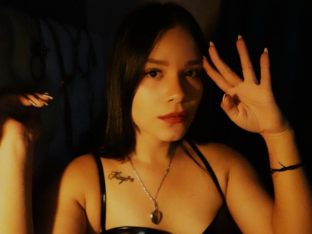SelenaRogue with dark hair, red lipstick, and black latex bra, showing off her tits with a silver necklace, making a peace sign with her hand, under warm lighting.