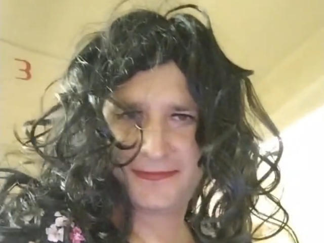 LeraSissy tiene una peluca rizada de color negro, un pequeño aro en la nariz y una ligera sonrisa con lápiz labial rojo. Tiene la piel pálida, sin senos ni genitales visibles. Ligeramente borroso, iluminación interior..