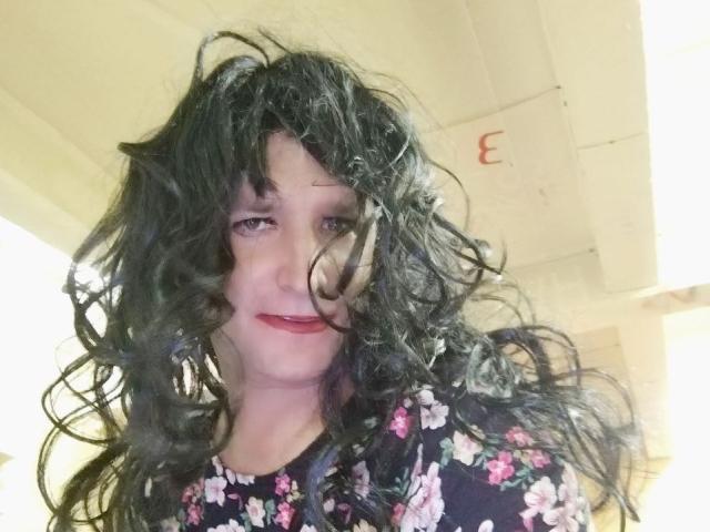 LeraSissy, com cabelos pretos cacheados e desalinhados, vestindo uma camisa florida, sorrindo levemente, com batom vermelho, pele clara, olhando para a câmera, em ambiente interno, sob iluminação fluorescente..