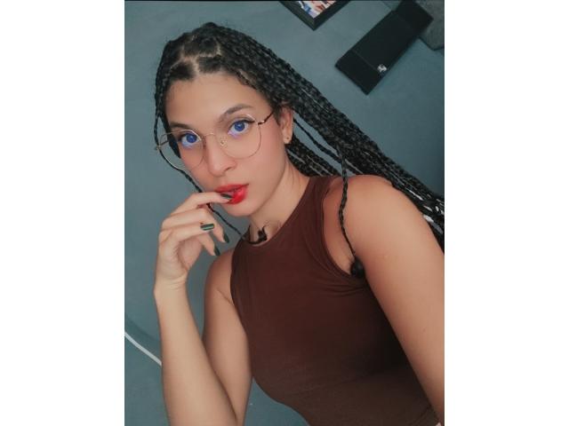 DylanyAllly69 com tranças longas, olhos azuis e lábios vermelhos, vestindo uma regata marrom, tocando seus lábios vermelhos com o dedo, usando óculos, em um selfie..
