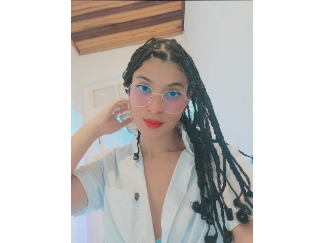 DylanyAllly69 with long black dreadlocks, wearing a white button-up shirt, pink round sunglasses, and red lipstick, touching her ear. Her tits are covered, but she's sexy.