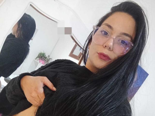 BiancaScott, avec de longs cheveux noirs, portant des lunettes et un pull noir, touchant ses cheveux, reflet dans un miroir, avec un anneau au nez et du rouge à lèvres rose. Ses fesses sont pixélisées..