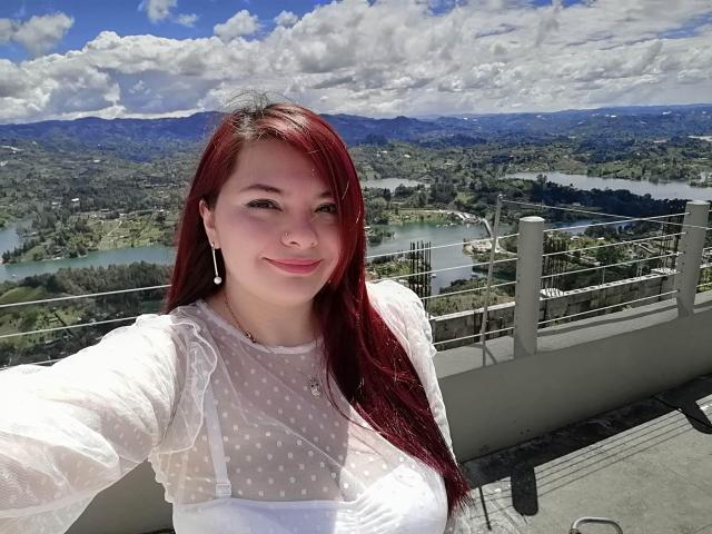 KatherinLatin with red hair, white polka-dot dress, small tits, smiling, earrings, on sunny balcony, mountains and lake in background, selfie, clear sky, fluffy clouds.