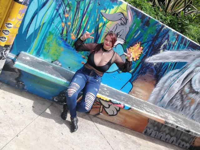 Katherin, une femme au physique voluptueux, avec de grandes seins, portant un haut noir transparent, un jean bleu déchiré et des bottes noires, assise devant des graffitis colorés, faisant un signe de la paix et souriant..