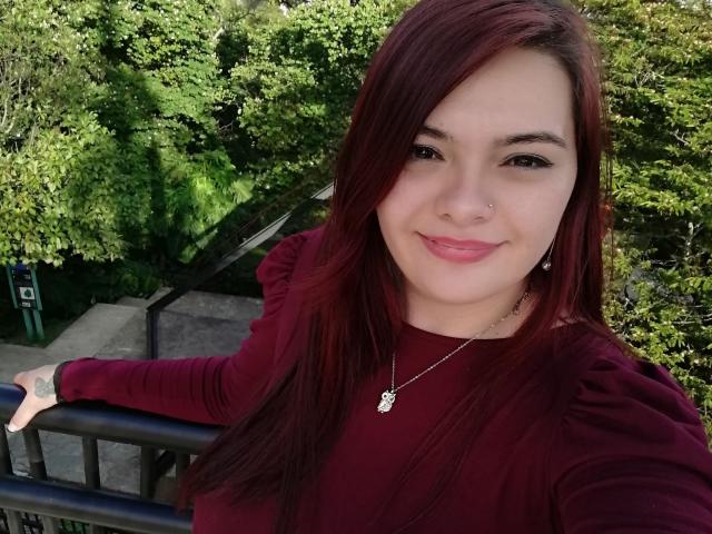 KatherinLatin with red hair, nose ring, and pink lipstick, wearing a maroon top, standing with a hand on a railing, smiling, necklace with a small pendant.