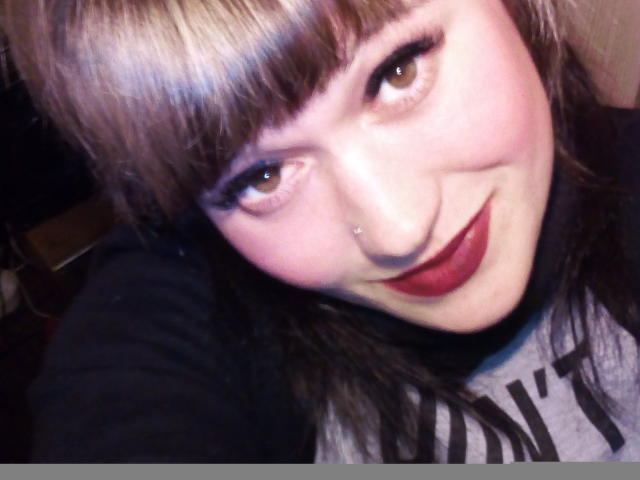 HollyLaBerry's face, showing her white skin, brown eyes, red lipstick, and nose piercing, with blonde-brown hair and black eyeliner, smiling at the camera.