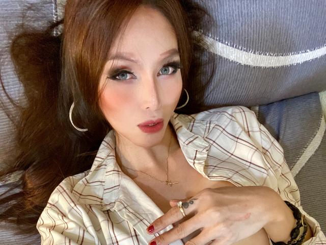 LaurentSuprema with brown hair, blue eyes, and pink lips, wearing a plaid shirt, hoop earrings, and a gold necklace, lying on a grey couch, hand on chest.