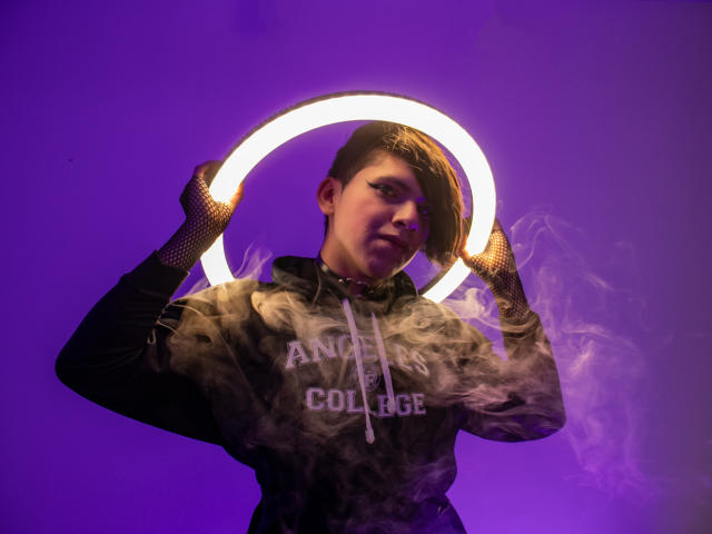 Saturni segurando uma luz de anel brilhante acima da cabeça, vestindo um moletom escuro e luvas de rede de pesca, com iluminação roxa e fumaça. Cabelo castanho curto, expressão confiante..