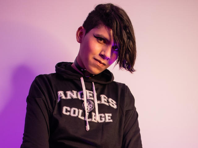 Saturni with short, dark hair, side-swept, wearing a black "Angels College" hoodie, looking at camera with subtle smile, purple and pink lighting on left side of face.