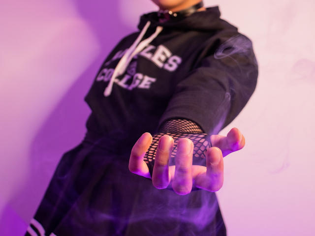 Saturni's black hoodie, fishnet glove, and outstretched hand with a cigarette, purple smoke, and choker, in a low-angle shot. Boobs not visible. Ass not visible. Pussy not visible.