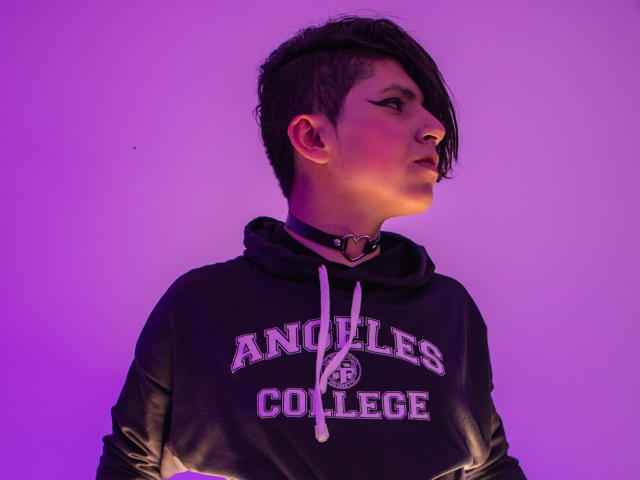 Saturni with short, dark, side-swept hair, wearing a black hood, choker with heart, and purple-lit background, looking to the right with a serious expression.