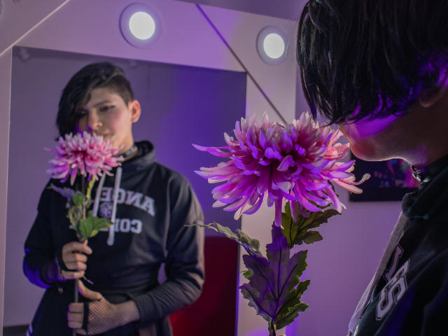 Saturni, con i capelli neri e corti, indossa una felpa nera e annusa un fiore di dahlia viola e rosa davanti a uno specchio. Il riflesso mostra Saturni che tiene un altro dahlia..