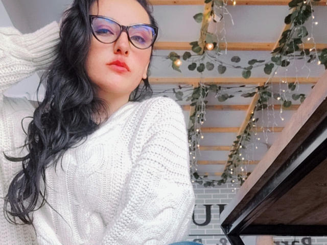Natalia, avec ses longs cheveux noirs, ses lunettes, son rouge à lèvres rouge et son pull en tricot blanc, regarde vers le bas avec une expression légèrement boudeuse, une main dans ses cheveux. Le plafond est orné de poutres en bois et de verdure..