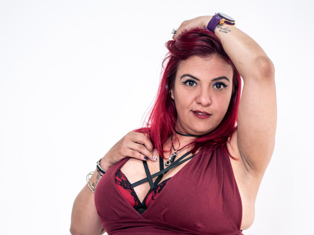 SammyOwens with red hair, wearing a maroon dress with black lace, exposing her big tits, arm raised, looking at camera, purple wristband, choker, and tattoos on her wrist.