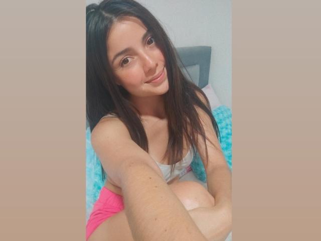 HeidyMillerr with long dark hair, smiling, wearing a gray top and pink shorts, holding her knee, showing her tits, sitting on a blue fluffy pillow.