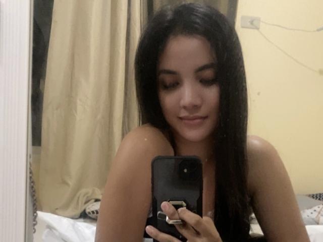 PeniaLove with dark hair, medium tits, holding phone, smiling, mirror selfie, bare shoulders, beige curtains, soft lighting, casual, cute, confident.