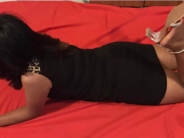 Cyah está acostada en una cama roja, vestida con un vestido negro que deja ver su trasero, con tacones altos rosas, aretes de estampado de leopardo y cabello negro..
