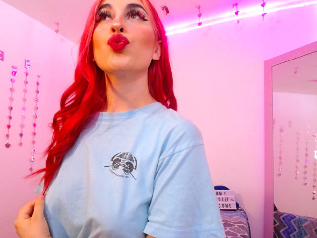 SammyViolet with bright red hair, pouty red lips, and a blue shirt, showing her tits. She's wearing long lashes, and her hand is on her hip.