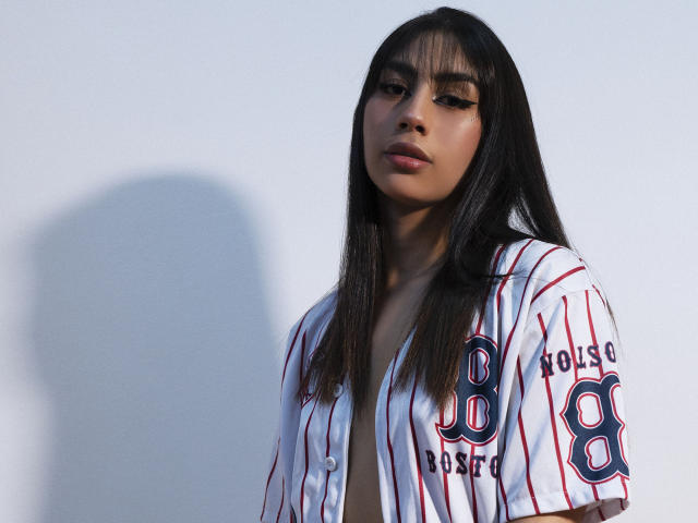 JessicaSoler with long black hair, wearing a baseball shirt with red and blue stripes, looking down with a serious expression, shadow on the wall. Boobs slightly visible under shirt.