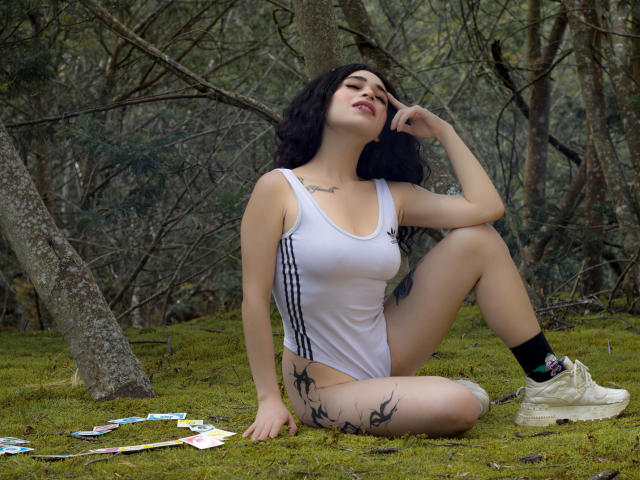 Hesttia with black curly hair, white tank top, black tattoos on thigh, sitting on moss, hand on head, white sneakers, black socks, forest background, looking up, red lipstick.