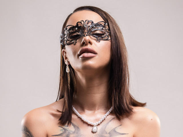 AwwLolla, avec sa coupe de cheveux courte marron, son masque en dentelle noire, son collier de perles et ses boucles d'oreilles, exhibe sa poitrine et ses tatouages, et a l'air séduisante avec ses lèvres légèrement entrouvertes..