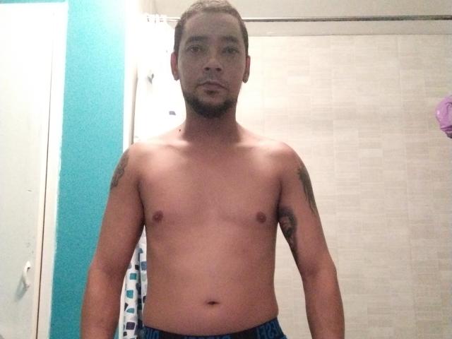 RocioHotMan, senza camicia, con piccoli capezzoli scuri e un tatuaggio sulla spalla destra, indossa pantaloni blu, è in piedi in un bagno e guarda direttamente la fotocamera..